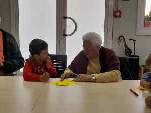 Àvia i nen fent manualitats junts a una taula blanca amb papers grocs i retoladors de colors en un centre cívic