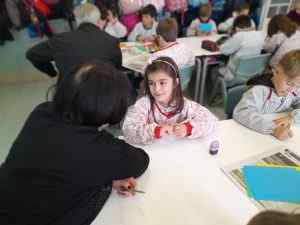 Nena somrient asseguda a una taula d'escola parlant amb una dona, amb altres estudiants treballant al fons de l'aula.