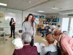 Jove professora ensenyant a un grup de persones grans en una aula amb pissarra i materials educatius.