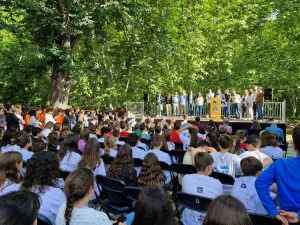 Estudiants asseguts escoltant una presentació a l'aire lliure amb professors dempeus en una plataforma sota arbres verds