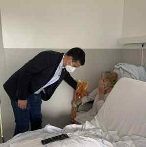 Home amb mascareta visitant dona hospitalitzada, oferint-li flors en una habitació d'hospital blanca
