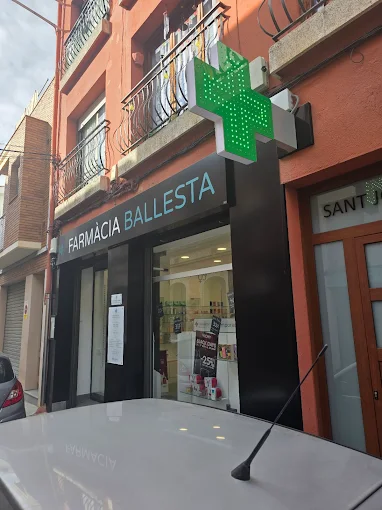 Farmàcia Ballesta amb creu verda il·luminada a la façana d'un edifici de maó vermell al carrer